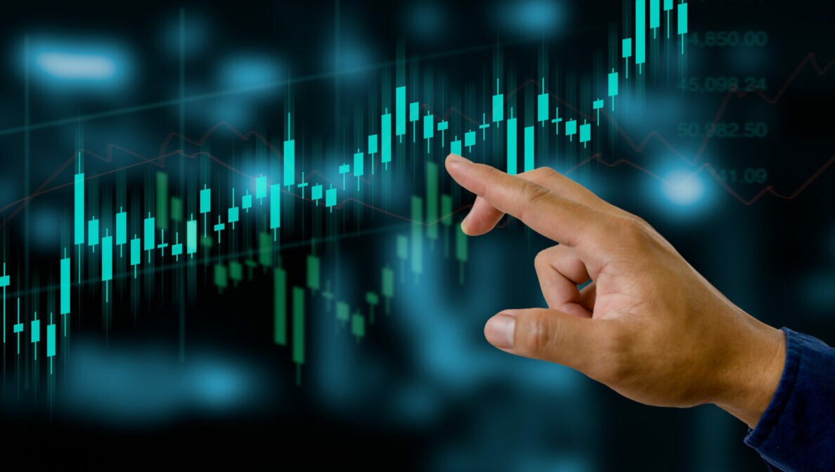 Eine Person zeigt mit dem Zeigefinger auf einen Bildschirm mit Kursverläufen und Kerzendiagrammen.