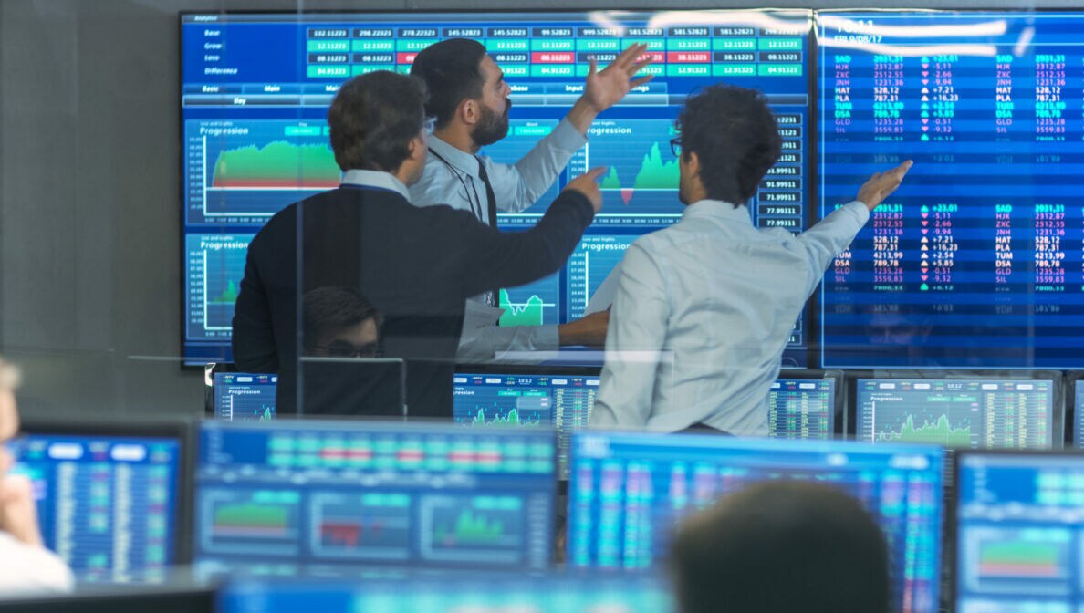 Mehrere Personen telefonieren in einem Raum mit Glasfenstern oder schauen auf Bildschirme, auf denen zahlen und Kursverläufe zu sehen sind.