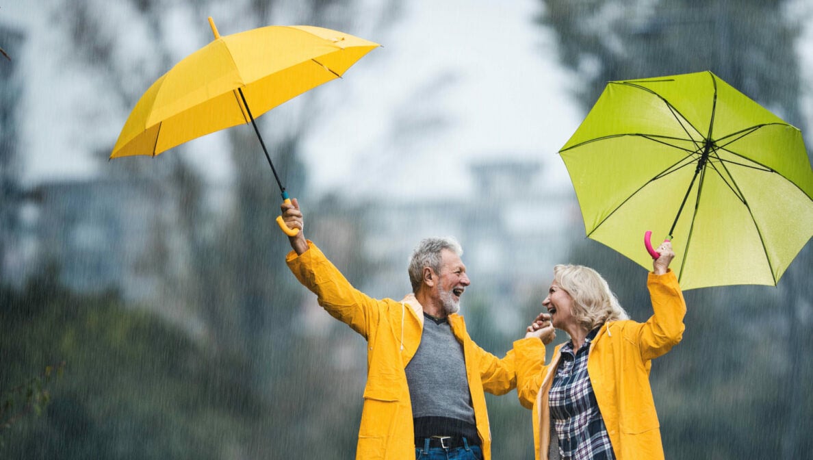 Zwei ältere Personen in gelben Regenmänteln halten im Regen ihre gelben Regenschirme in die Höhe und schauen sich lachend an.
