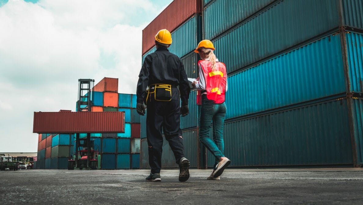 Zwei Personen in Arbeitskleidung und gelben Schutzhelmen gehen neben gestapelten Containern entlang. Im Hintergrund hebt ein Kran einen Container.