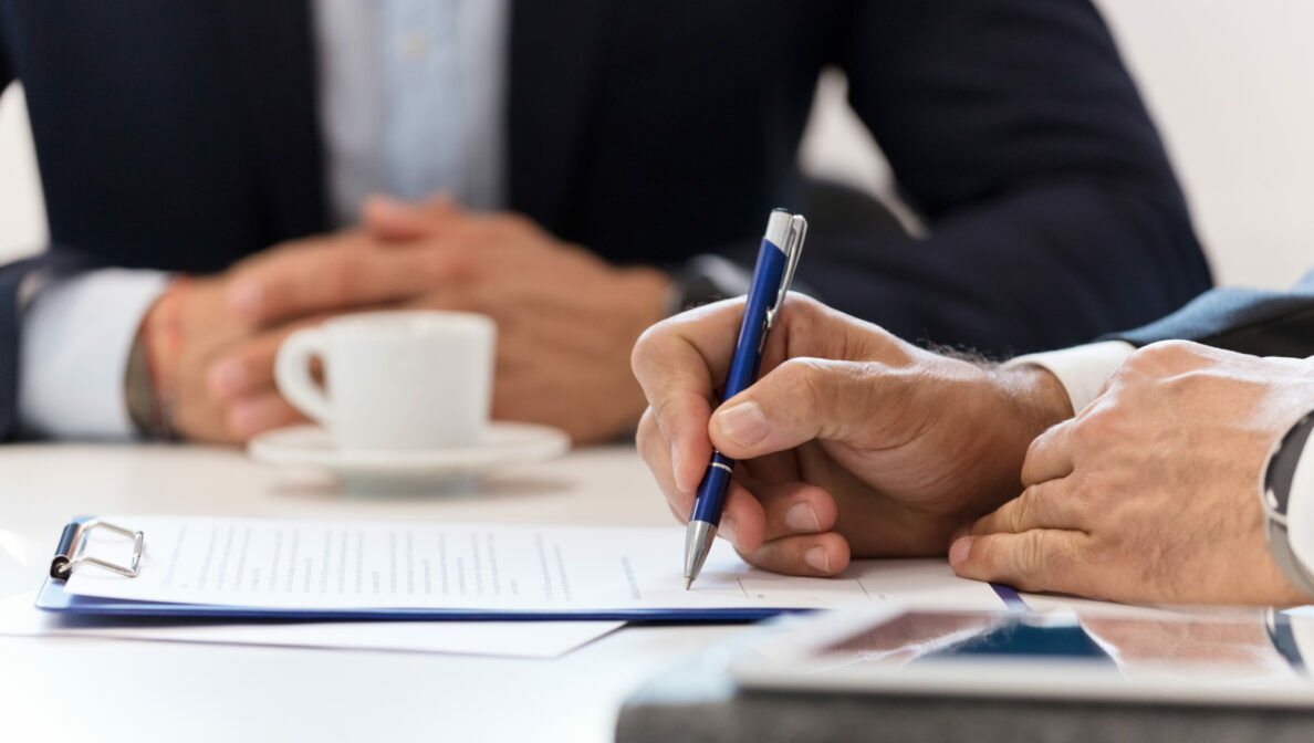 Zwei Personen im Anzug sitzen an einem Tisch mit Espressotasse und einem Dokument an einem Klemmbrett. Eine Person schreibt mit einem Kugelschreiber auf das Dokument.