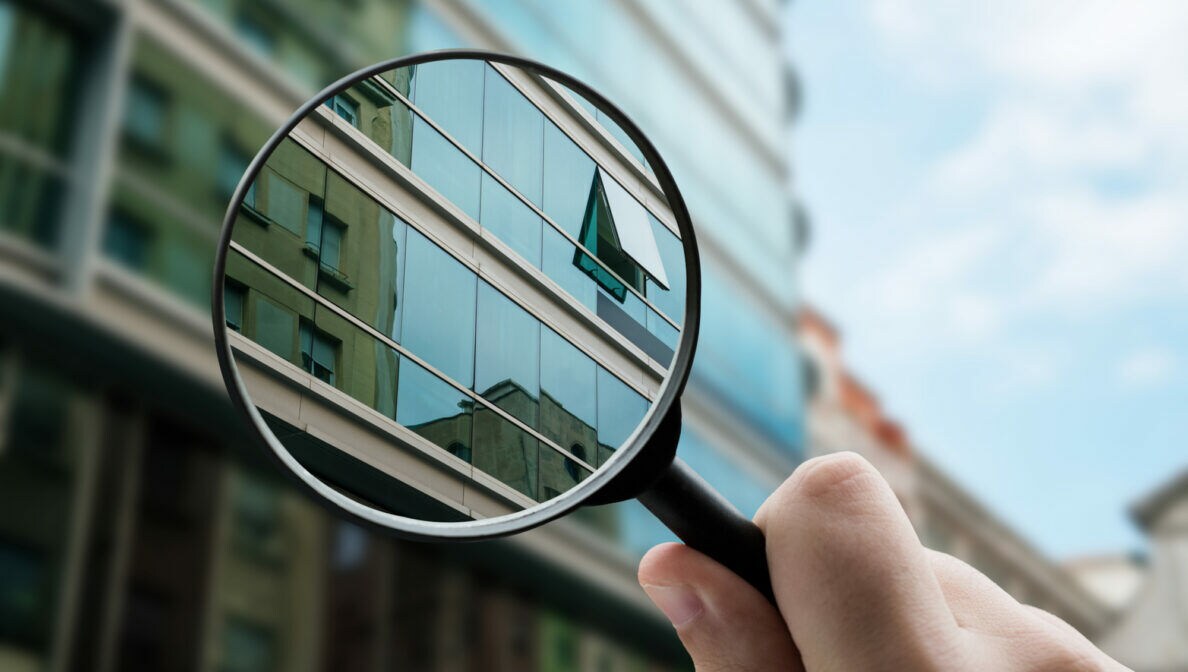 Eine Person hält eine Lupe, durch die der vergrößerte Ausschnitt eines gläsernen Hauses zu sehen ist, dessen weitere Fassade im Hintergrund verschwommen zu erkennen ist.
