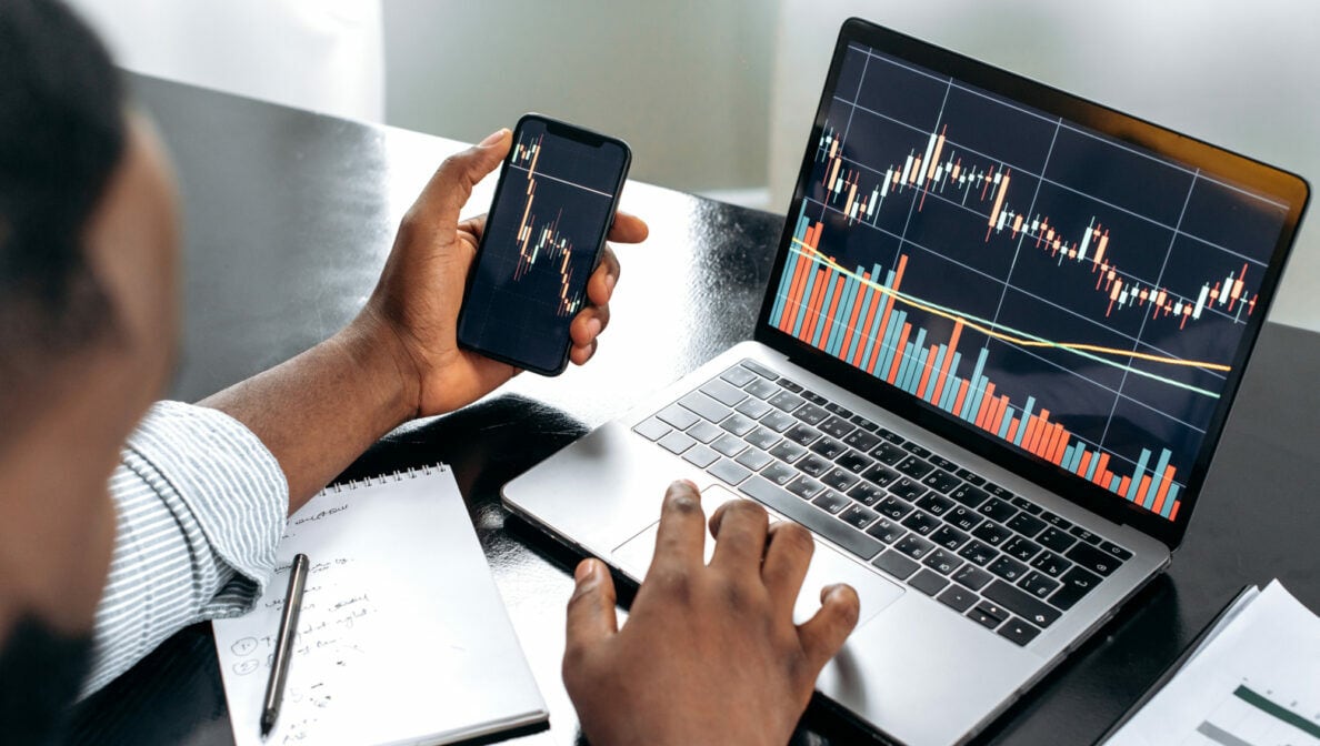 Eine Person hält ein Smartphone in der Hand. Auf dem Bildschirm ist ein Aktienkurs zu sehen. Im Hintergrund ein Notebook, das ebenfalls Kurse zeigt.