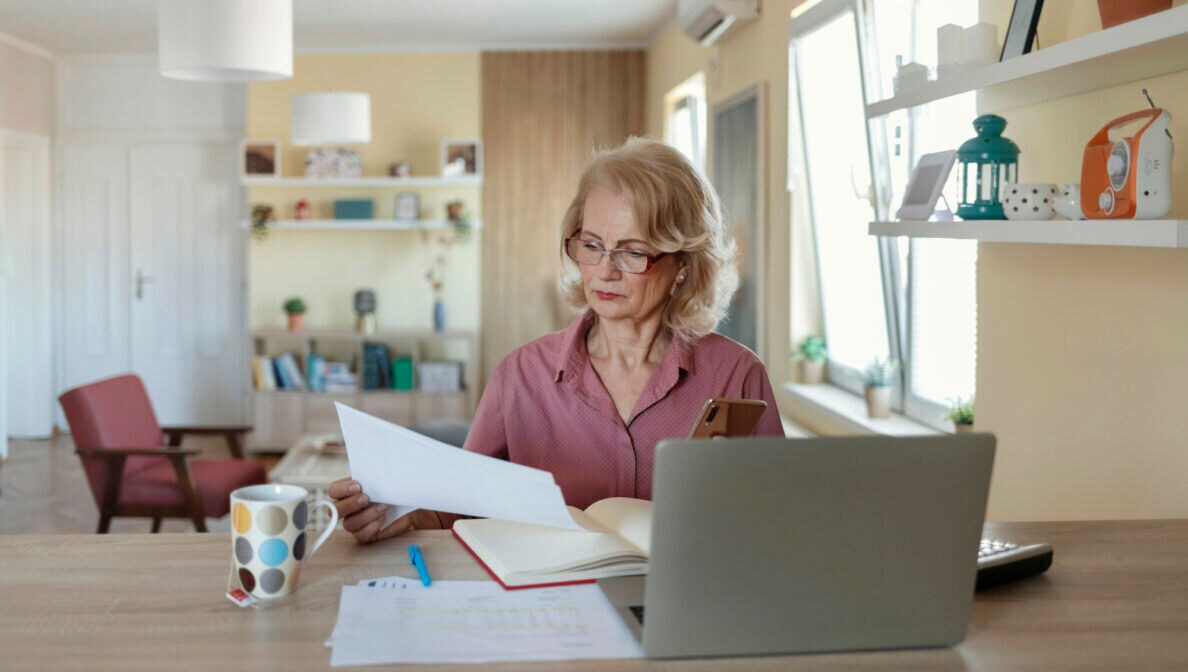Eine Person sitzt an einem Tisch vor einem aufgeklappten Laptop, einem aufgeschlagenem Buch, einem Taschenrechner sowie einer Tasse mit Teebeutel und hält ein Blatt Papier in der Hand.