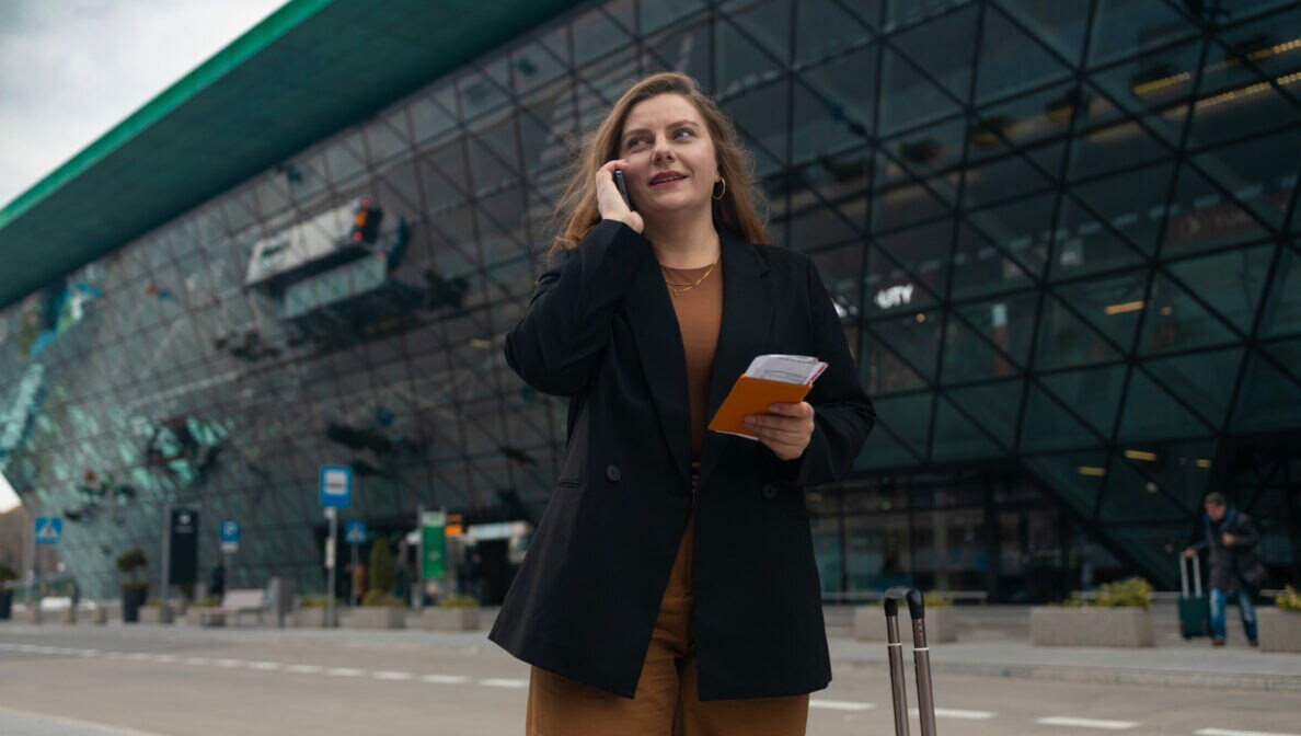 Eine Frau vor einem verglasten, modernen Gebäude hält mit einer Hand ein Telefon ans Ohr, in der anderen hält sie Reisedokumente. Neben ihr steht ein Koffer.