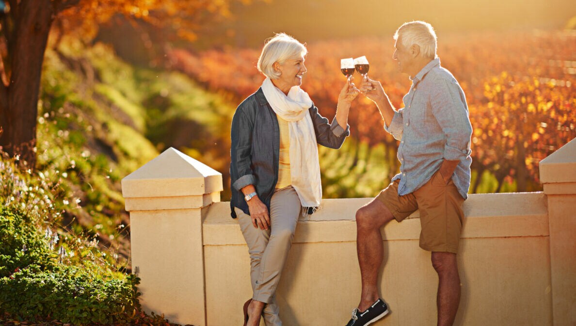 Ein älterer Mann und eine ältere Frau stoßen an eine Mauer gelehnt vor herbstlichen Laubbäumen mit einem Glas Rotwein an.