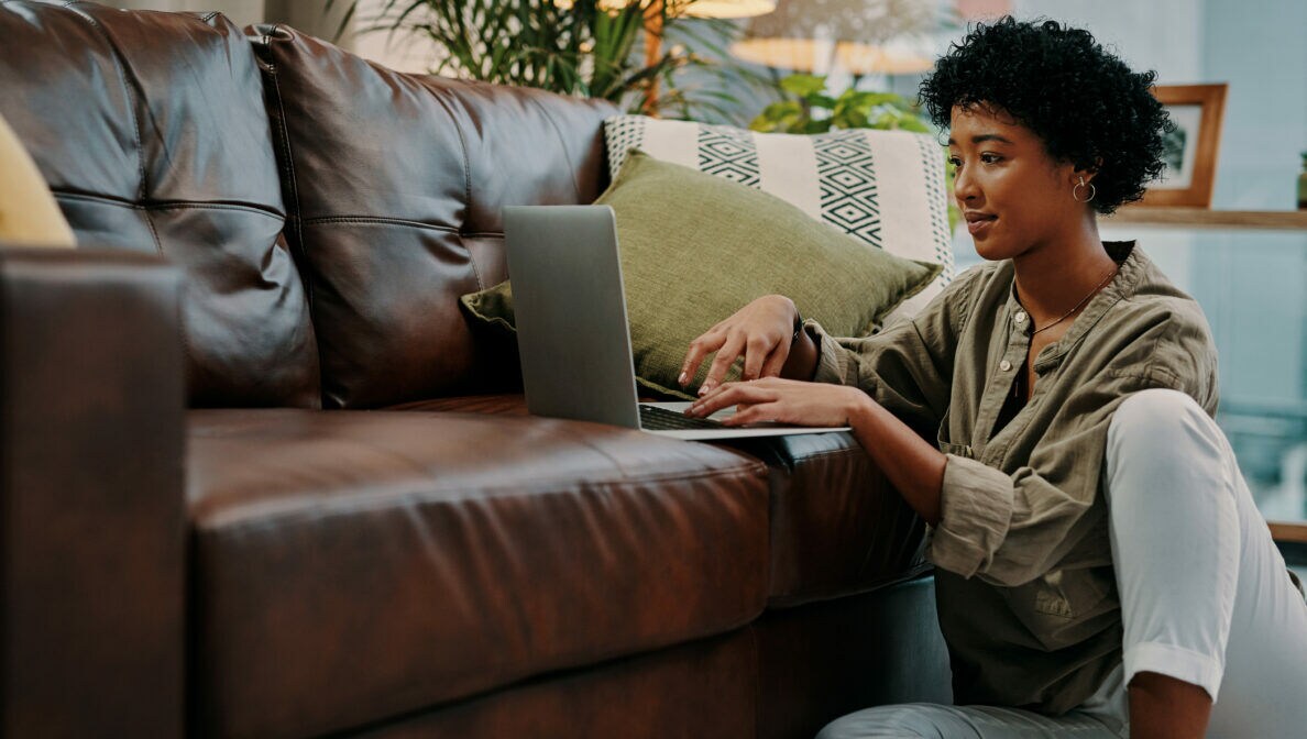 Eine junge Frau sitzt auf dem Boden neben dem Sofa und benutzt ein Notebook.