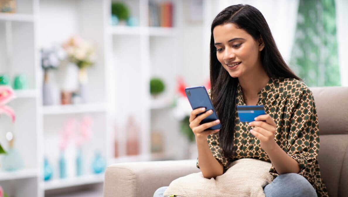 Eine Person sitzt im Schneidersitzt auf einem Sofa und hält sowohl eine Kreditkarte als auch ein Smartphone in den Händen.