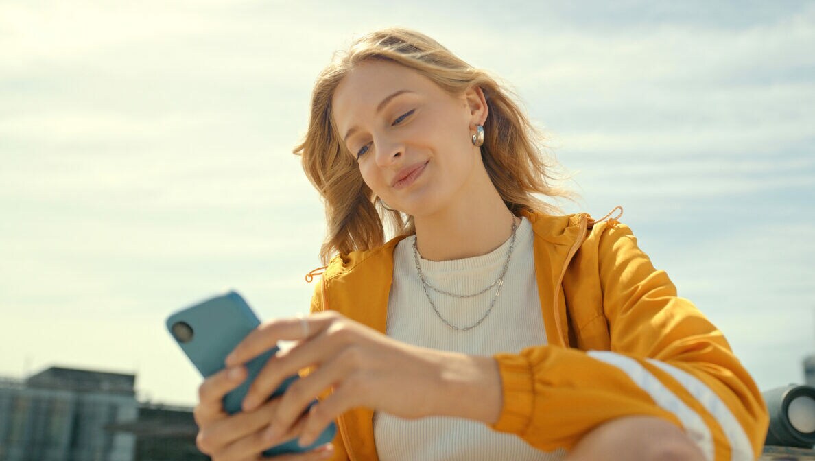 Eine junge Person sitzt im Freien und hält ein Smartphone in ihren Händen.
