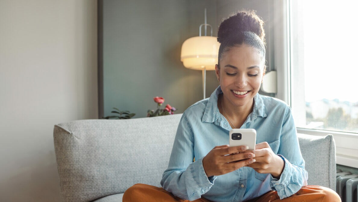 Eine Person sitzt im Schneidersitz auf einem Sofa und blickt auf ihr Smartphone, das sie in beiden Händen hält.