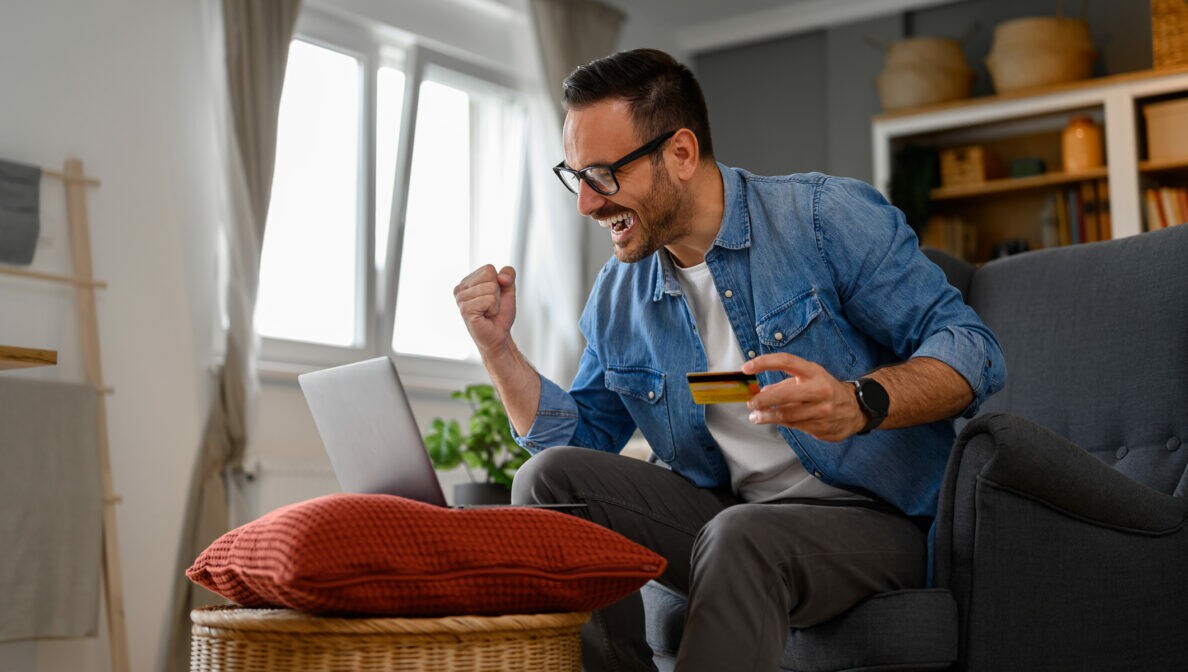 Ein Mann sitzt auf einem Sofa vor einem aufgeklappten Laptop, der auf einem Hocker steht. In einer Hand hält er eine Geldkarte, die andere ballt er zu einer Faust.