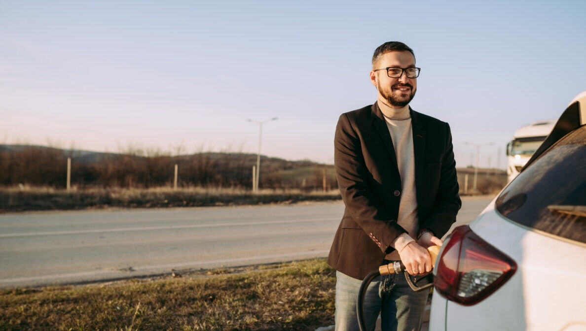Eine Person steht vor ihrem Auto und hält den Zapfhahn fest, der im Auto steckt.