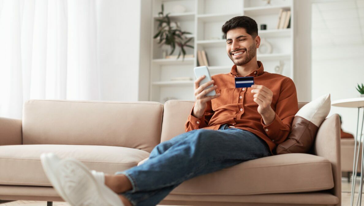 Eine Person sitzt mit ausgestreckten Beinen auf einem Sofa und hält eine Kreditkarte sowie ein Smartphone in den Händen.