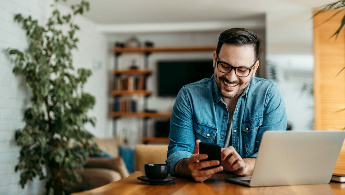 Eine Person sitzt an einem Tisch und blickt auf ein Smartphone, das sie in den Händen hält. Vor ihr stehen ein geöffnetes Laptop und eine Kaffeetasse.
