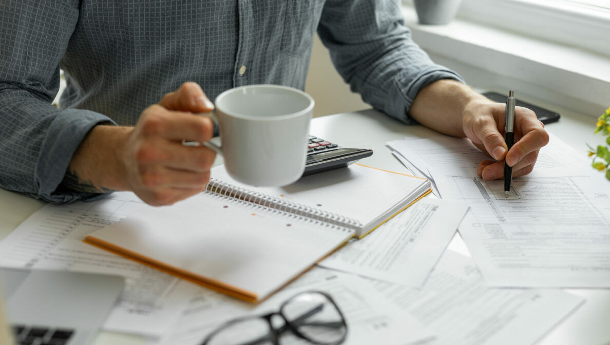 Eine Person am Tisch hält eine Tasse und einen Kugelschreiber in Händen. Auf dem Tisch liegen Dokumente, ein Taschenrechner und eine Brille.