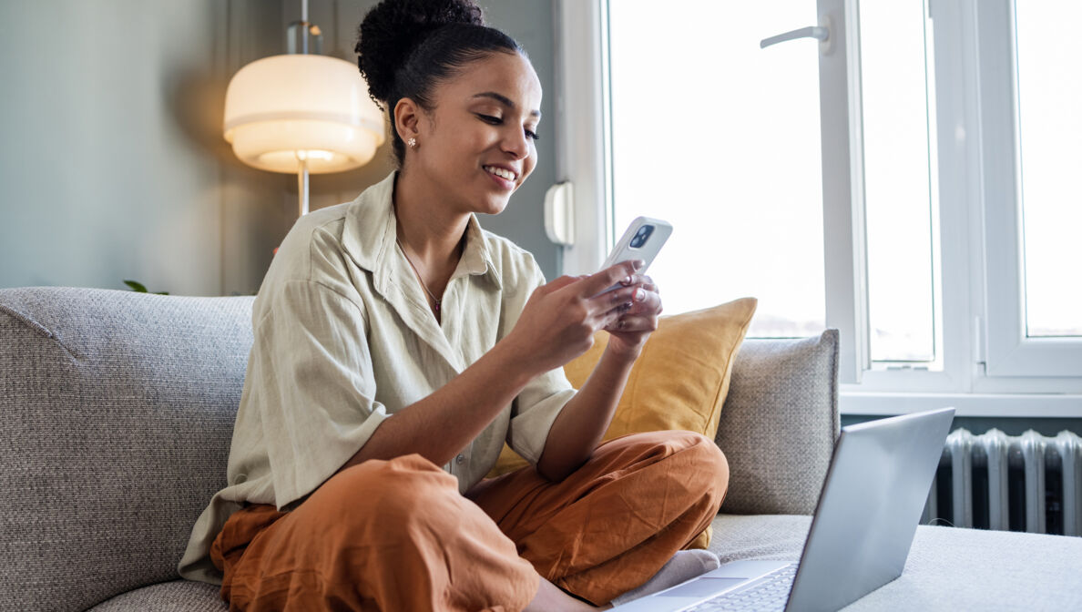 : Eine Frau sitzt im Schneidersitz auf einer Couch und schaut lächelnd auf ihr Smartphone. Vor ihr steht ein Laptop.
