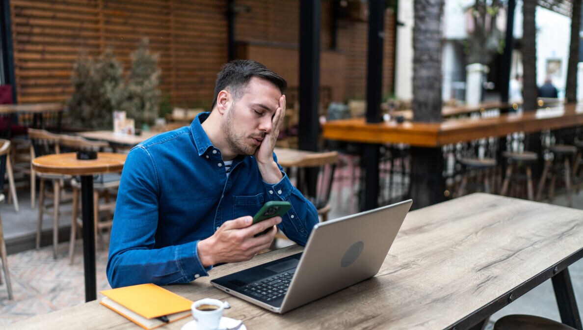 Ein Mann sitzt an einem Tisch vor einem Laptop und hält ein Smartphone in der Hand. Er hat die Augen geschlossen und legt eine Hand über sein Gesicht.