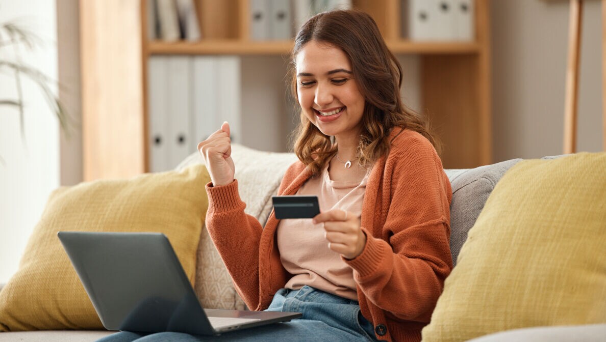Eine Person sitzt lächelnd auf einem Sofa, einen Laptop auf den Beinen und eine Kreditkarte in der Hand und ballt eine Faust.