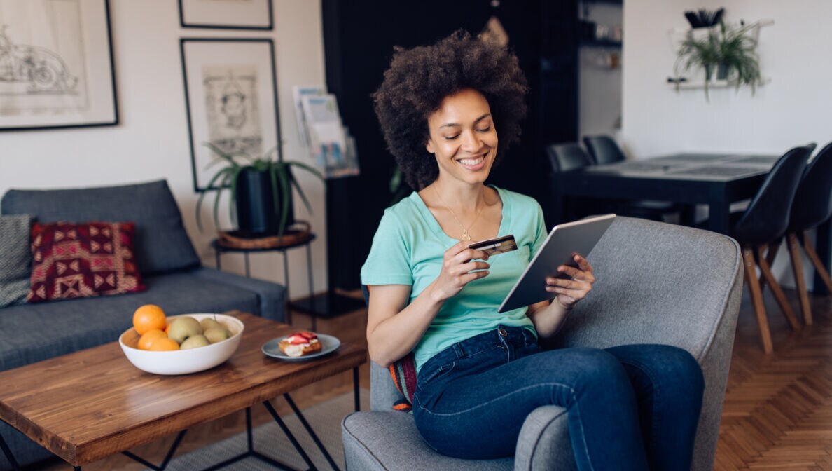 Person sitzt auf einem Sessel und schaut auf ihr Tablet und auf ihre Kreditkarte