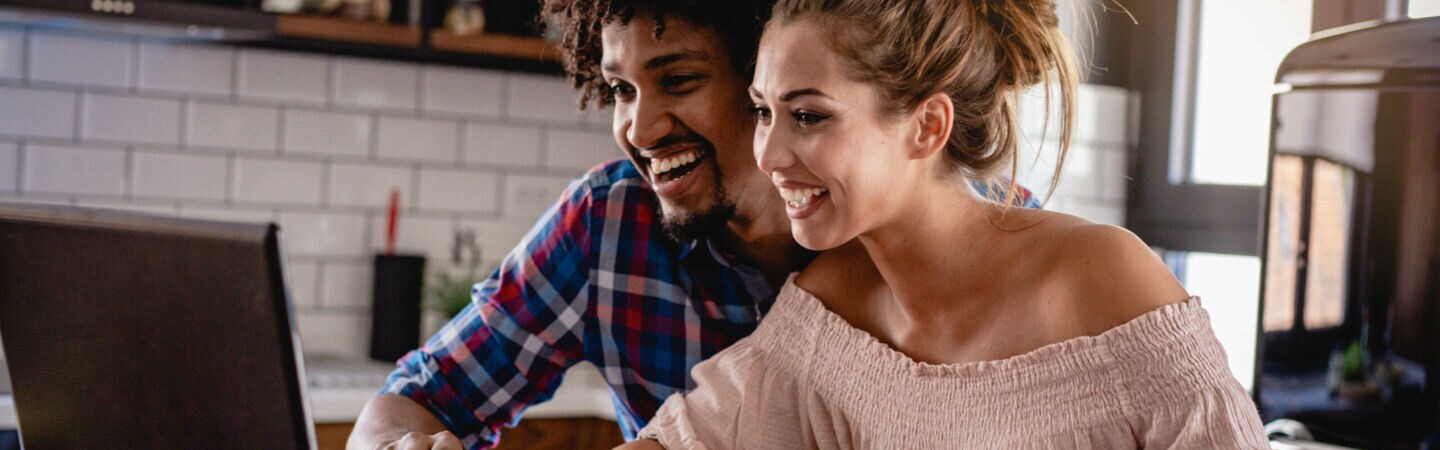 Ein junges, lächelndes Paar vor einem Laptop, Taschenrechner und Zetteln am Küchentisch