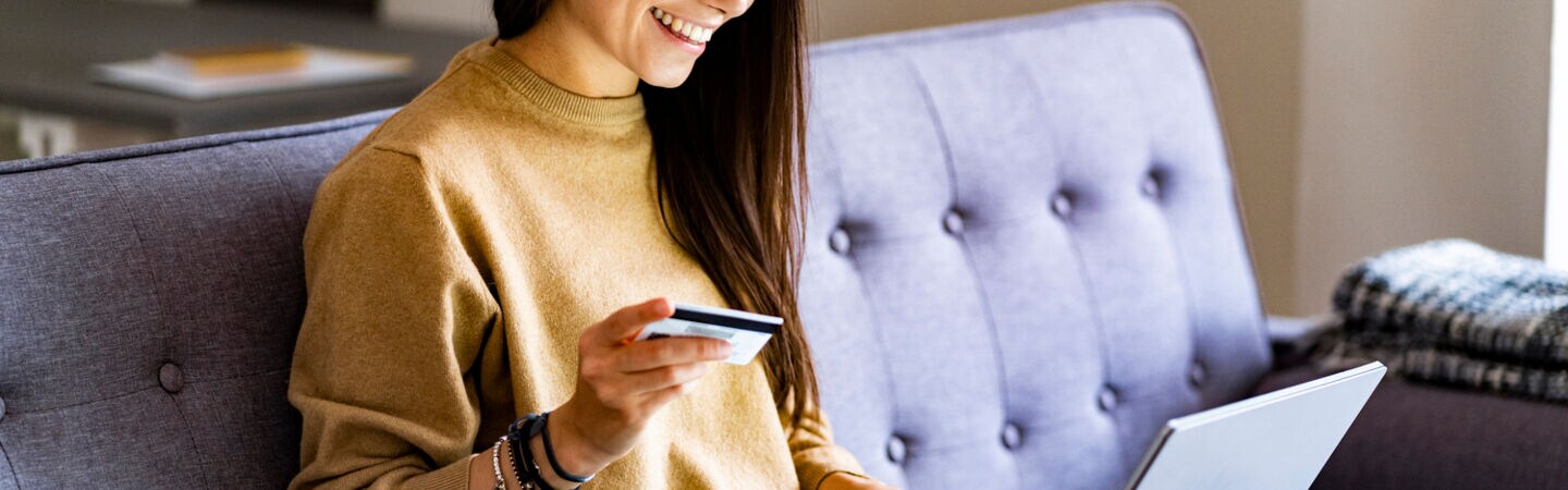 Eine Frau im Schneidersitz auf einem Sofa mit Laptop im Schoß und Kreditkarte in der Hand