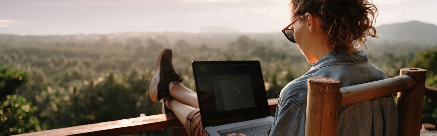 Eine Frau sitzt im Freien auf einer Terrasse, arbeitet an einem Laptop mit Blick ins Grüne.