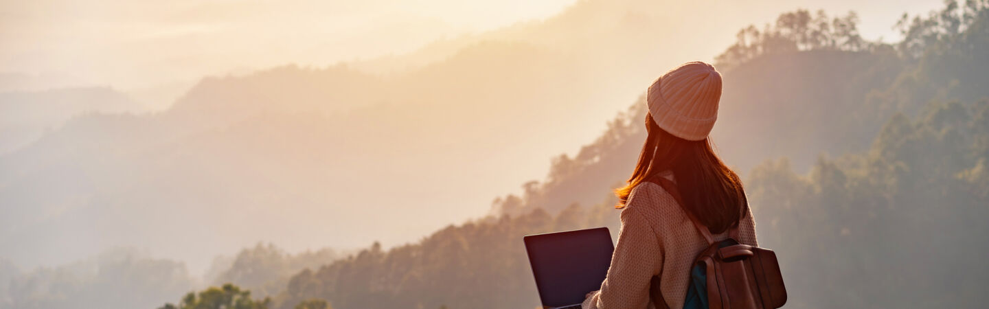 Eine Person mit Laptop sitzt auf einem hölzernen Untergrund und blickt auf ein Naturpanorama mit Bergen und Sonnenaufgang