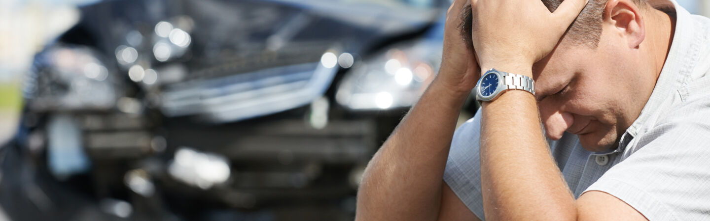 Eine Person hockt am Straßenrand und hat den Kopf in die Hände gestützt, im Hintergrund steht ein kaputtes Auto