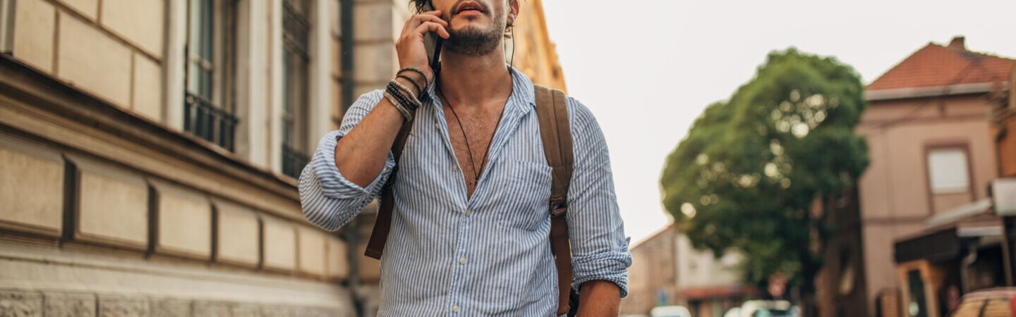 Mann mit Bart, Sonnenbrille und Rucksack auf dem Rücken steht auf einem Bürgersteig und telefoniert