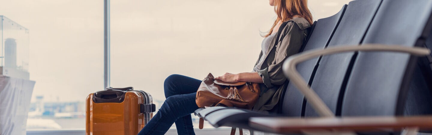 Eine Person sitzt mit einem Koffer im Flughafen und blickt aus dem Fenster, an dem gerade ein Flugzeug vorbeifliegt