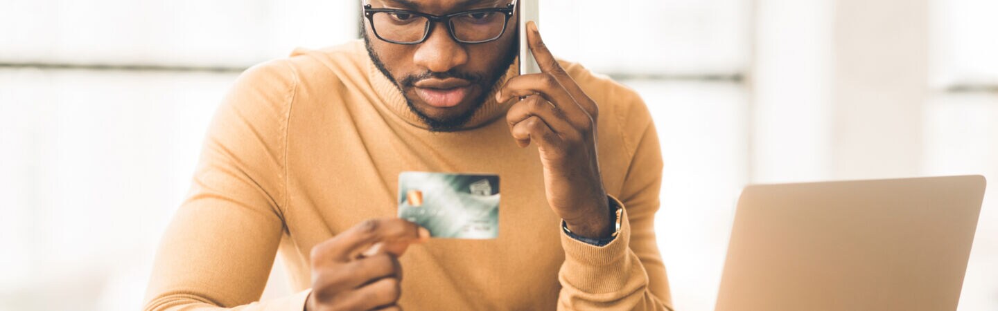 Eine Person sitzt telefonierend vor einem geöffneten Laptop am Tisch und blickt auf seine Bankkarte in der Hand.