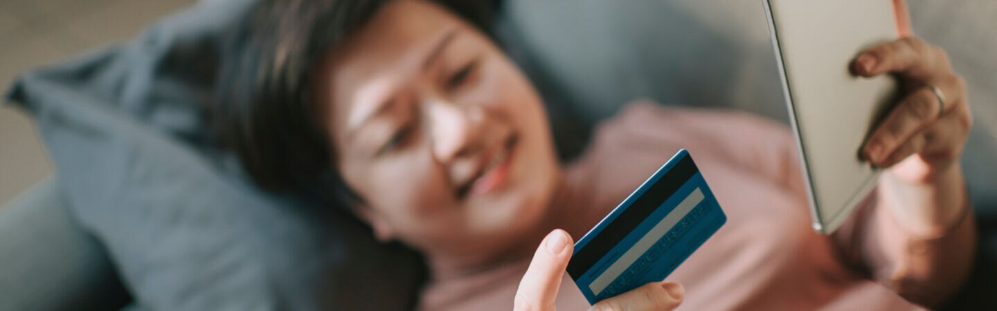 Eine Person liegt auf der Couch. Sie hält in der einen Hand eine Kreditkarte und in der anderen ein Smartphone.