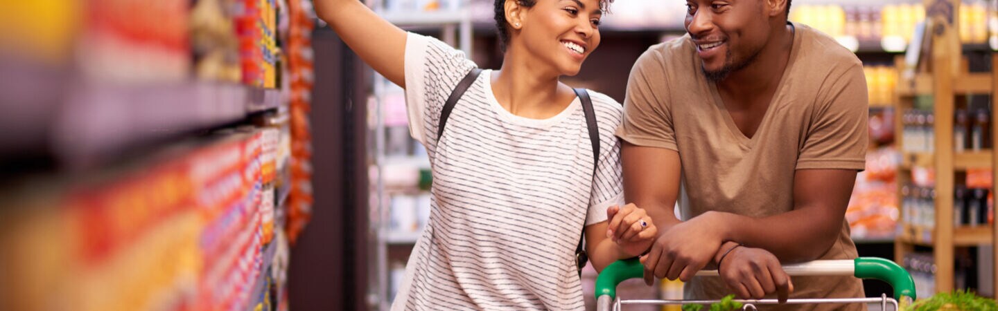 Zwei Personen zwischen Supermarktregalen. Sie schieben einen mit Gemüse gefüllten Einkaufswagen.