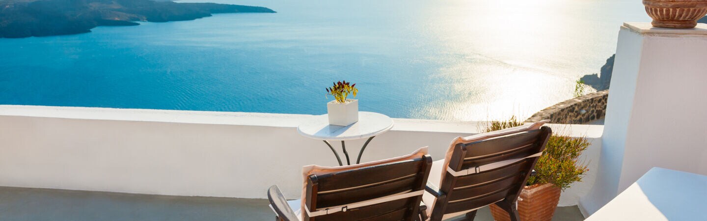 Zwei Holzstühle stehen auf einer sonnigen Terrasse mit Blick übers Meer.