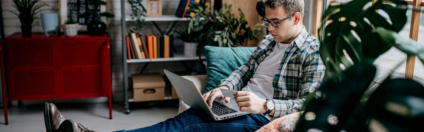 Ein Mann sitzt auf dem Sofa, die Füße hochgelegt. Er hat ein geöffnetes Notebook auf dem Schoß und eine Karte in der Hand.