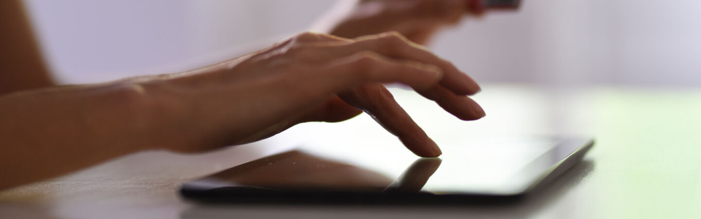 Nahaufnahme einer Hand, die auf einem Tablet tippt. Im Hintergrund ist eine zweite Hand zu erkenne, die eine Bankkarte in der Hand hält.