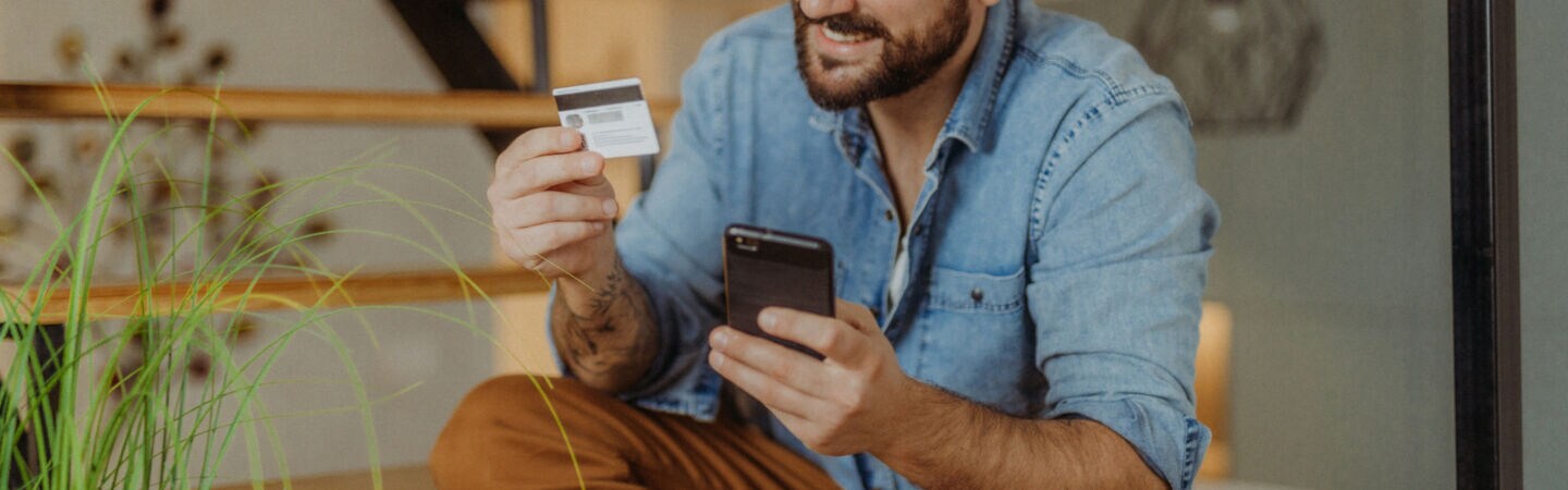 Ein Mann sitzt lächelnd auf den Stufen einer Treppe. In der einen Hand hält er eine Kreditkarte, auf die er schaut. In der anderen Hand hält er ein Smartphone