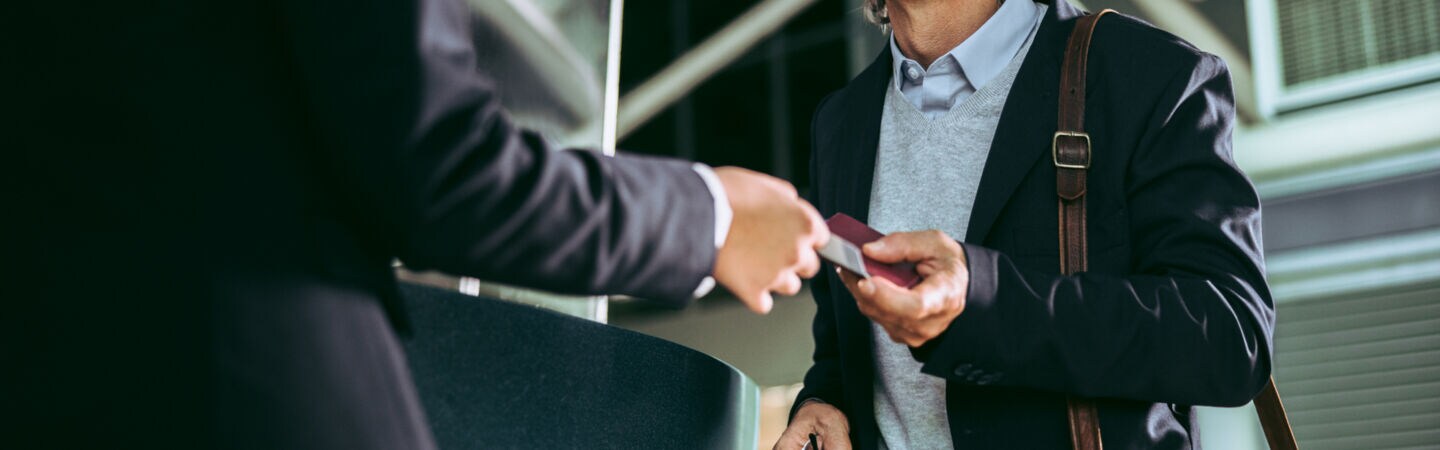 Ein Mann steht lächelnd vor einem Tresen und bekommt von einer Silhouette in schwarz einen Reisepass in die Hand gedrückt