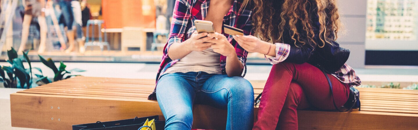 Zwei Personen sitzen in einer Shoppingmall auf einer Bank und schauen gemeinsam auf ein Smartphone und eine Kreditkarte.