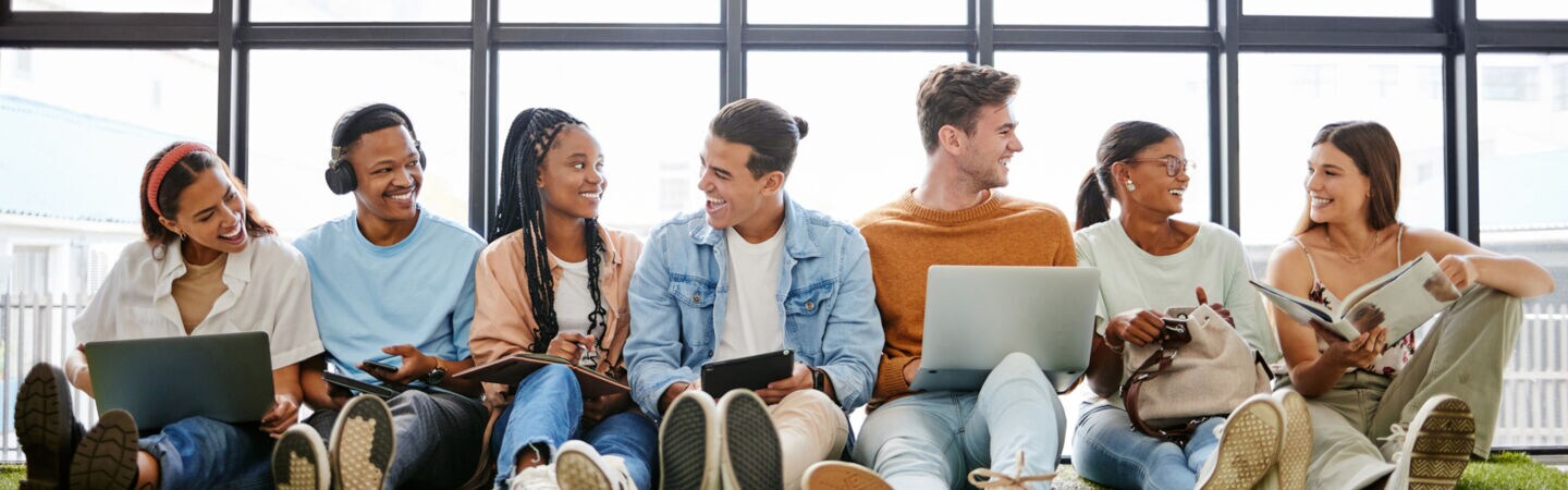 Mehrere junge Personen sitzen nebeneinander auf dem Fußboden an eine Fensterfront gelehnt.