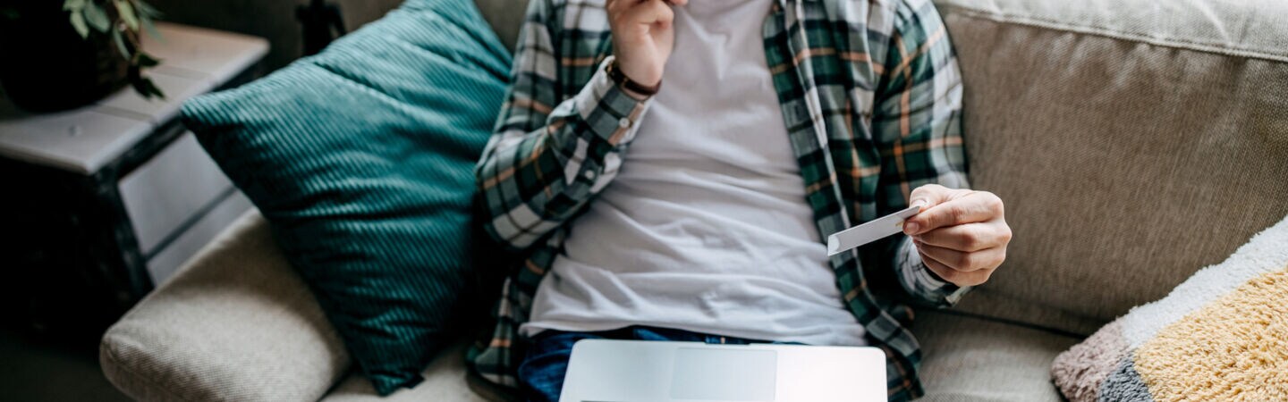 Eine Person aus der Aufsicht, die telefonierend auf einem Sofa sitzt, eine Bankkarte in der Hand hält und ein Laptop auf dem Schoß liegen hat.