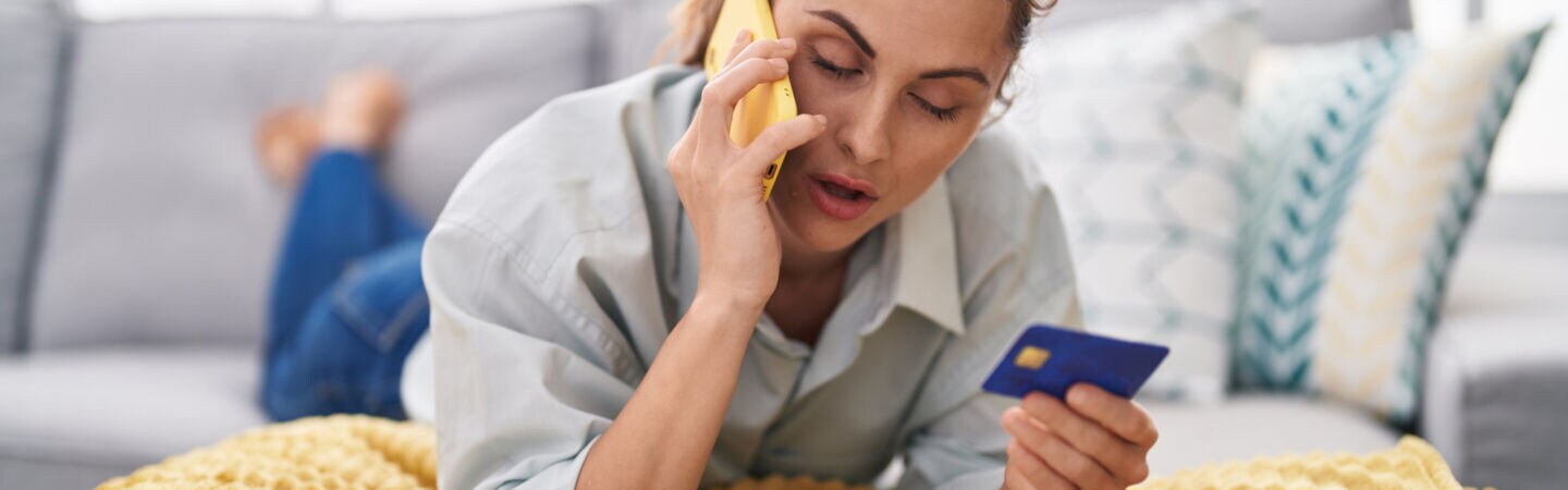 Eine Person liegt bäuchlings und telefonierend auf dem Sofa und hält eine Kreditkarte in der Hand.