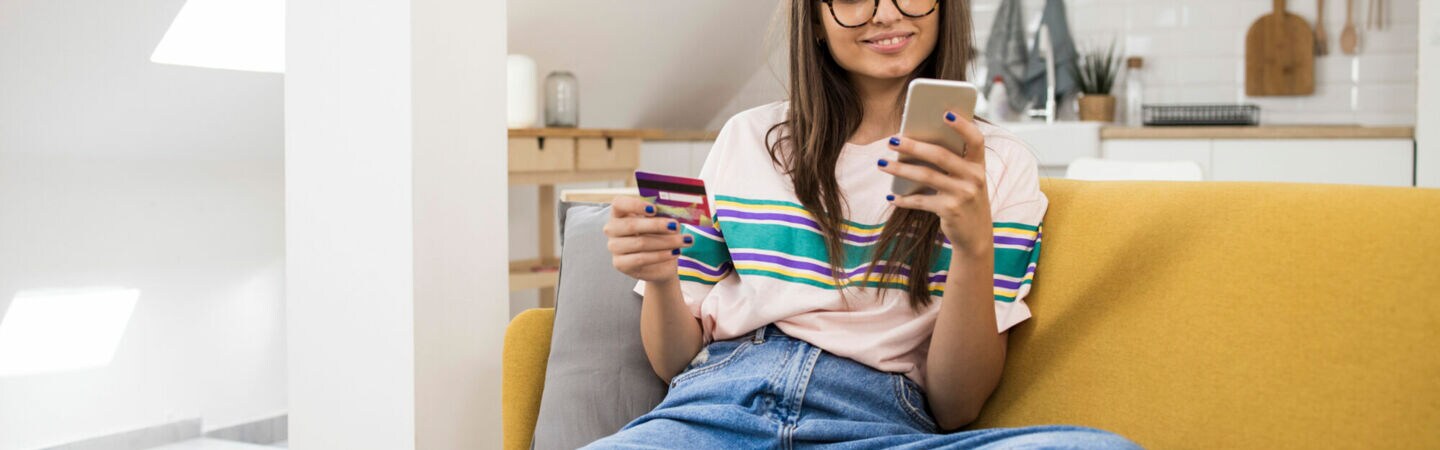 Eine Jugendliche mit langen braunen Haaren und Brille sitzt auf einer Couch und schaut lächelnd auf ein Smartphone in ihrer rechten Hand. In der linken Hand hält sie eine Bankkarte.
