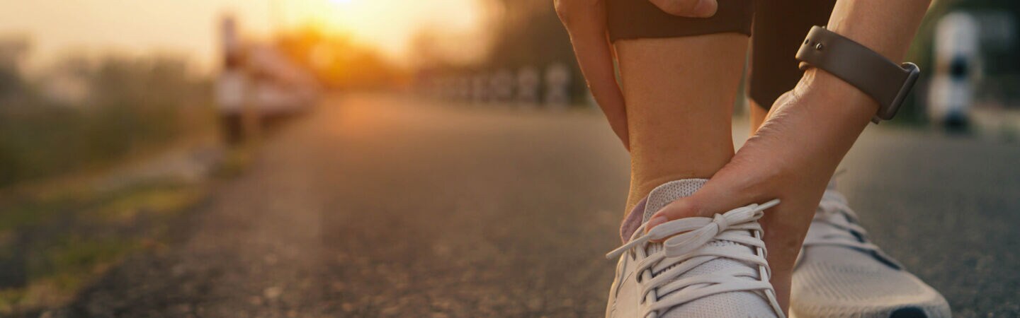 Eine Person in Laufschuhen hockt auf einem asphaltierten Weg und hält sich mit beiden Händen den Knöchel.