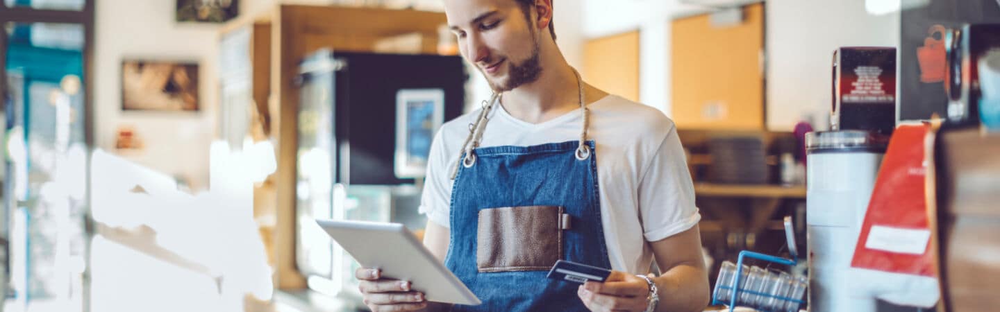 Ein junger Barista steht in einem Café, er hat eine Schürze an und hält in der einen Hand ein Tablet und in der anderen Hand eine Kreditkarte