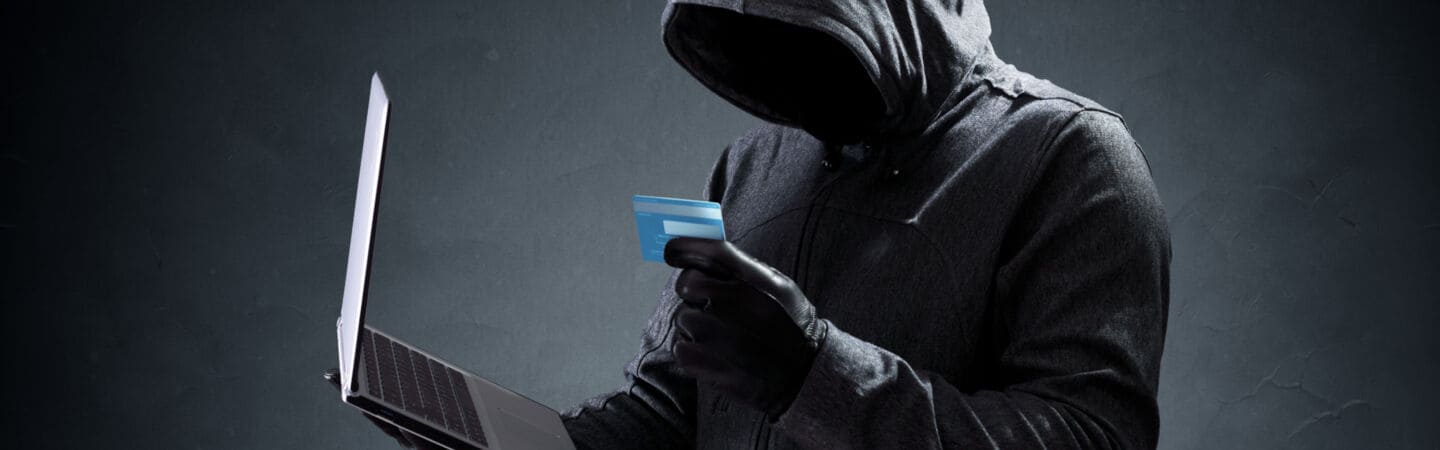 Eine Person im Hoodie, die Kapuze über den Kopf gezogen, hält in den schwarz behandschuhten Händen ein Notebook und eine Kreditkarte