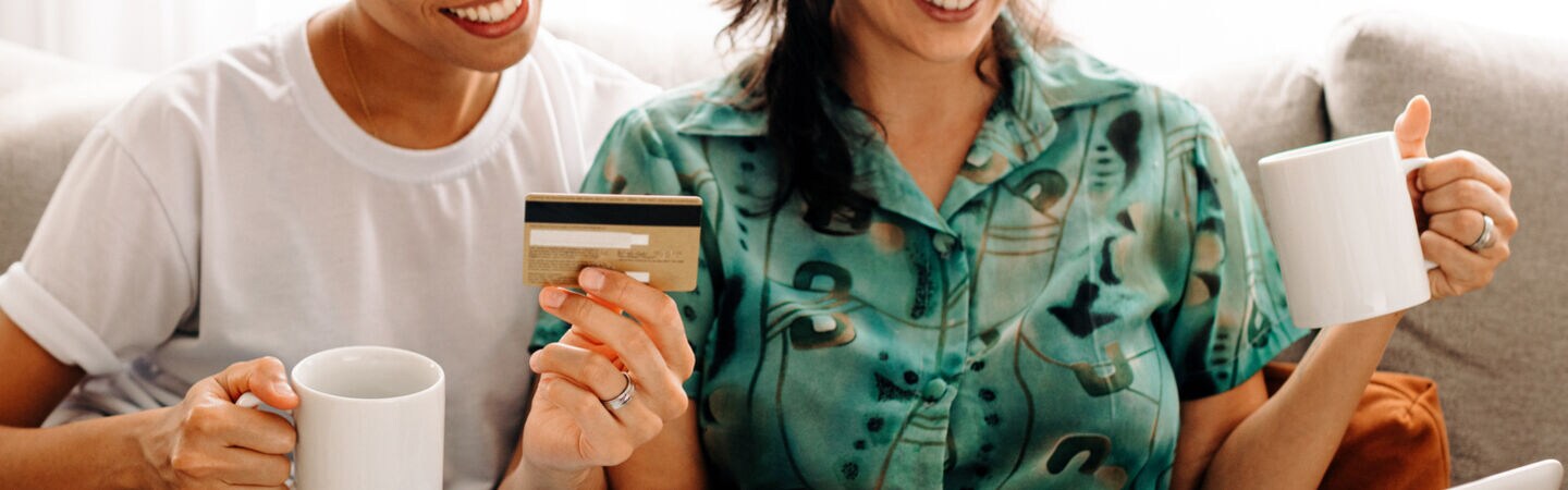 Zwei auf dem Sofa sitzende Personen halten eine Tasse und eine Kreditkarte in der Hand. Eine Person hat einen Laptop auf dem Schoß.