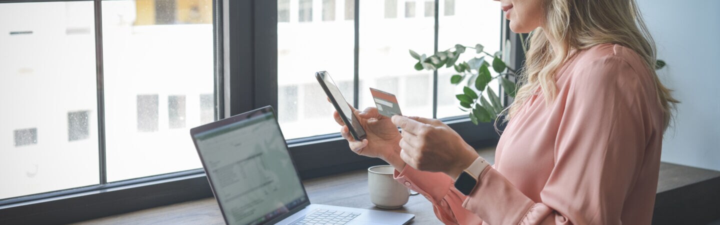 Eine Frau steht mit Handy und Kreditkarte in der Hand vor einem Tisch, auf dem ein Laptop steht