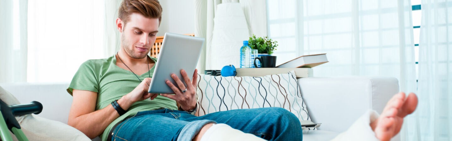 Eine Person mit gebrochenem Bein sitzt auf einem Sofa und schaut auf sein Tablet, neben ihr stehen Krücken.