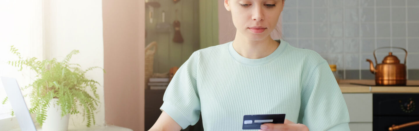Eine Person sitzt am Küchentisch, schaut auf ihre Bankkarte und hat mehrere Dokumente vor sich liegen sowie die andere Hand auf einem geöffneten Laptop.